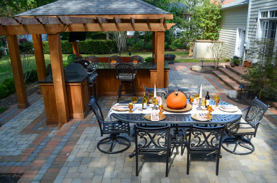 Outdoor dining table with pumpkins and glassware in front of wood pavilion, paver patio, and covered kitchen.